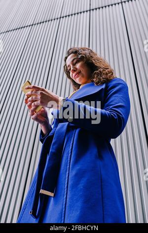 Donna d'affari in trench cappotto utilizzando il telefono cellulare mentre si sta in piedi parte anteriore della parete grigia Foto Stock