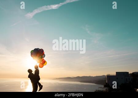 Uomo in silhouette che porta figlia con palloncini sul mare Foto Stock