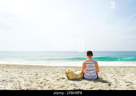 Vista posteriore dell'uomo e del suo cane seduto sulla spiaggia guardando le onde dell'oceano in una giornata di sole limpida. Animali da compagnia maschi caucasici pastore canino, vista mare, sabbia, wa Foto Stock