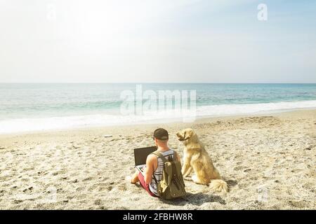 Vista posteriore dell'uomo e del suo cane seduto in spiaggia guardando le onde dell'oceano, giorno di sole chiaro. Programmatore FIT freelance maschio che lavora su computer portatile in mare w Foto Stock