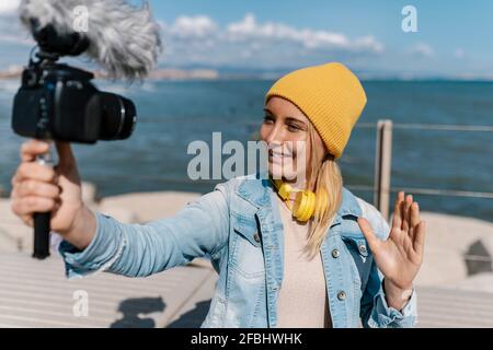 Donna sorridente con cappello a maglia che svita la mano mentre vlogging attraverso fotocamera durante il fine settimana Foto Stock