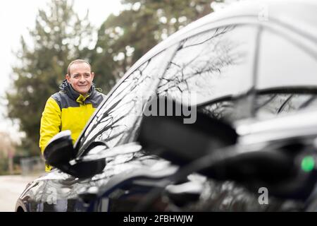 Uomo sorridente in piedi davanti all'auto elettrica Foto Stock