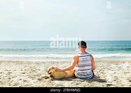 Vista posteriore dell'uomo e del suo cane seduto sulla spiaggia guardando le onde dell'oceano in una giornata di sole limpida. Animali da compagnia maschi caucasici pastore canino, vista mare, sabbia, wa Foto Stock