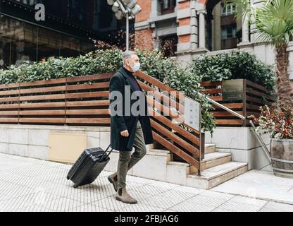 Uomo maturo che indossa una maschera protettiva che cammina con i bagagli sentiero Foto Stock