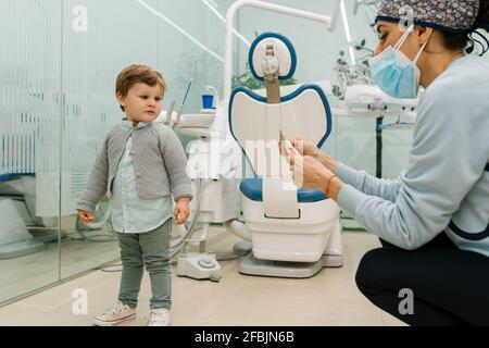 Bambino che guarda la dentista pediatrica femminile con il viso protettivo maschera in clinica Foto Stock