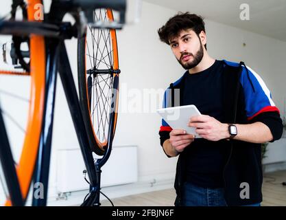 Uomo che guarda la bicicletta upturnt mentre tiene il tablet digitale a. casa Foto Stock