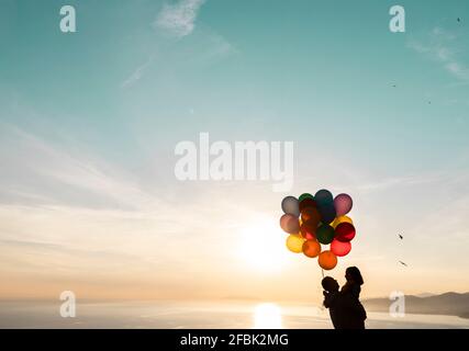 Uomo che porta la figlia che tiene palloncini colorati durante il tramonto Foto Stock