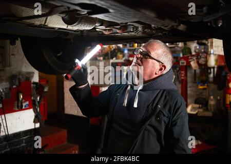 Meccanico maschile senior con torcia che esamina l'auto in officina di riparazione Foto Stock