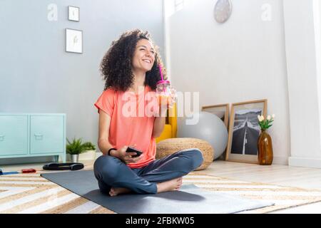 Donna allegra che ha succo seduto con il telefono cellulare sul tappetino in soggiorno Foto Stock