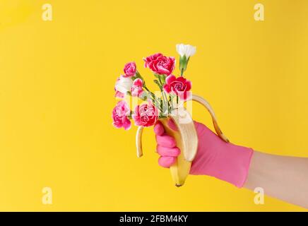 Arrangiamento creativo con mano femminile in guanto rosa e banana frutta con bouquet con fiori colorati su sfondo giallo brillante. Minimo conce Foto Stock