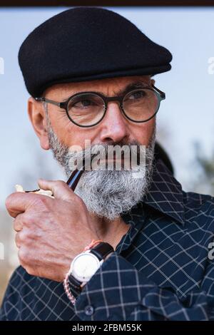 Uomo bearded in cappello piatto che guarda via mentre tiene fumare tubo Foto Stock