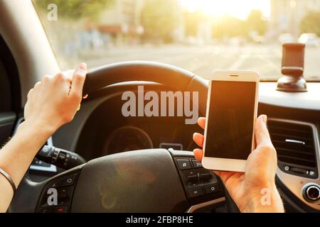 Giovane donna che tiene il telefono delle cellule dello schermo vuoto mentre guida l'automobile. Mano del conducente femminile sul volante e controllo del suo smartphone in un veicolo in movimento. D Foto Stock