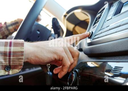 Giovane uomo in camicia a scacchi Hipster premendo i pulsanti sul pannello multimediale dell'auto, commutando la manopola della stazione radio. Primo piano della mano maschio sul veicolo Foto Stock