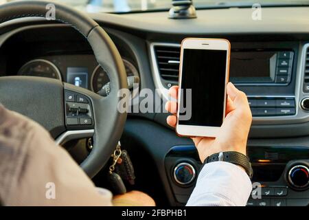 Giovane uomo che tiene il telefono delle cellule dello schermo vuoto mentre guida l'automobile. Mano maschile del conducente sul volante e controllo del suo smartphone in un veicolo in movimento. Non Foto Stock