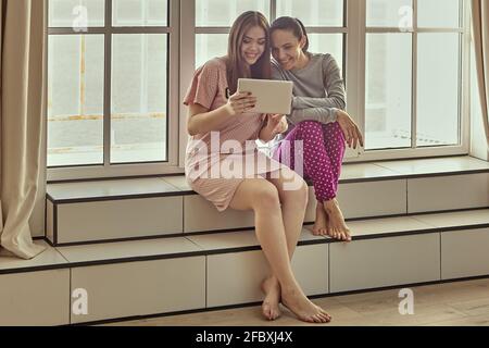 Due donne in sleepwear sono sedute sul davanzale e guardano video divertenti sullo schermo del tablet pc. Foto Stock