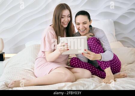 Madre con una figlia adulta sta guardando video divertente sullo schermo del computer tablet mentre si siede a letto. Foto Stock