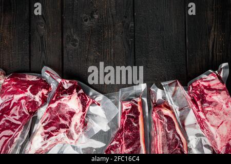 Carne di manzo cruda nel set di impacchi sottovuoto, tomahawk, t bone, bistecca di club, costolette e tagli di filetto, su sfondo di tavola di legno nero, fla vista dall'alto Foto Stock