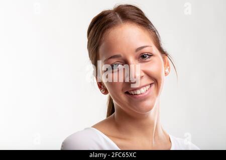 Bella giovane donna con i suoi capelli in una coda di ponytail e. un sorriso dolce e genuino guardando la fotocamera in un primo piano del ritratto della testa contro il retro di uno studio bianco Foto Stock