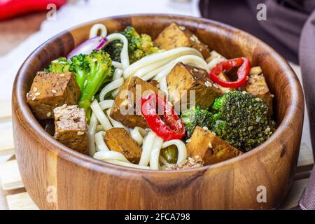 Tofu fritto vegetariano fatto in casa con tagliatelle e verdure (broccoli, pepe e cipolle) nella ciotola di legno su fondo cucina chiaro. Foto Stock