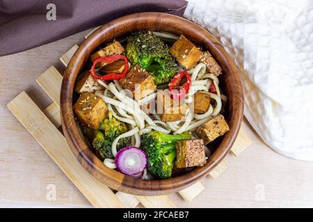 Vista dall'alto del tofu fritto vegetariano fatto in casa con tagliatelle e verdure (broccoli, pepe e cipolle) nella ciotola di legno sullo sfondo della cucina leggera Foto Stock