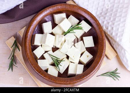 Vista dall'alto dei cubetti di tofu vegetariani biologici in ciotola rotonda con rosmarino fresco su sfondo di legno. Foto Stock