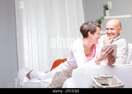 Coppia di mezza età che gode di una buona risata sui media sul loro tablet pc che si guardano l'un l'altro con la trasmissione via ir di sorrisi rilassati su un divano a casa Foto Stock