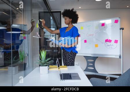 Gara mista businesswoman attaccando note memo su parete di vetro con lavagna bianca in background Foto Stock