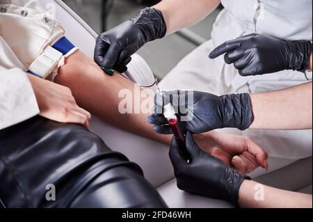 Chiudere la mano del medico con guanti neri che tengono la provetta con il sangue mentre l'assistente inserisce l'ago nel braccio del paziente. Scienziati di laboratorio medico che raccolgono campioni di sangue da giovane donna. Foto Stock