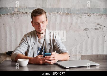 Vista frontale dello studente maschile che riposa con smartphone e una tazza di caffè dopo il lavoro sul computer portatile. Il giovane uomo in pullover grigio è seduto alla scrivania con il telefono nelle mani. Concetto di lavoro remoto. Foto Stock
