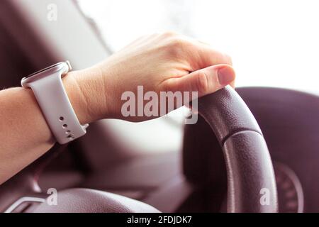 La mano di una donna con un orologio bianco sta tenendo in primo piano il volante di un'auto nera. Traffico su strada. Concetto di lezioni di guida. Abitacolo, speedomete Foto Stock