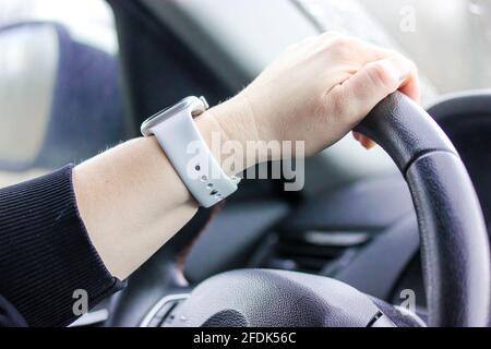 La mano di una donna con un orologio bianco sta tenendo in primo piano il volante di un'auto nera. Traffico su strada. Concetto di lezioni di guida. Abitacolo, speedomete Foto Stock