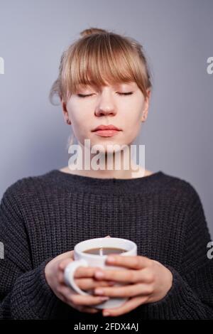 Concetto di pensiero sognante o positivo. Bella donna bionda caucasica in maglione nero che tiene una tazza di caffè o tè con gli occhi chiusi su sfondo grigio. Atmosfera romantica o caffè del mattino Foto Stock