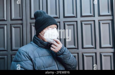 L'uomo più anziano in cappotto blu e cappello tiene il suo bianco maschera al suo viso all'aperto Foto Stock