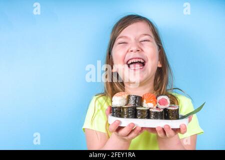 Carina bambina ama mangiare sushi. Allegra divertente caucasica bambina prescolare bionda con vari sushi roll e chopsticks. Su sfondo blu brillante Foto Stock