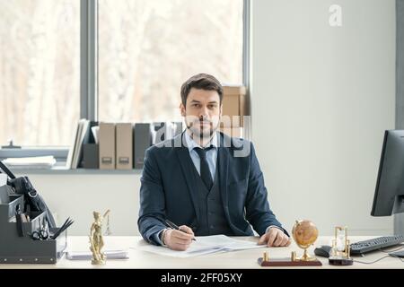Ritratto del soddisfare handsome avvocato bearded in vestito seduto a. scrivania ed esame di documenti in ufficio Foto Stock