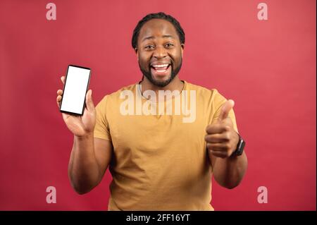 L'uomo afroamericano soddisfatto tiene uno smartphone con uno schermo vuoto e mostra il pollice in su, allegro ragazzo skinned scuro che fa pubblicità alla nuova app mobile isolata su sfondo rosso, concetto di mockup Foto Stock