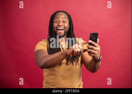 Sorprendo sorridendo uomo scuro skinned con lunghi Dreadlock che tengono il telefono, stupito ragazzo africano che legge buone notizie nel messaggio, guardando la macchina fotografica, scorrendo le notizie del feed, su sfondo rosso Foto Stock