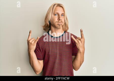 Giovane caucasico con capelli lunghi facendo gesto rock soffia guance con viso divertente. Bocca gonfia con aria, cattura aria. Foto Stock