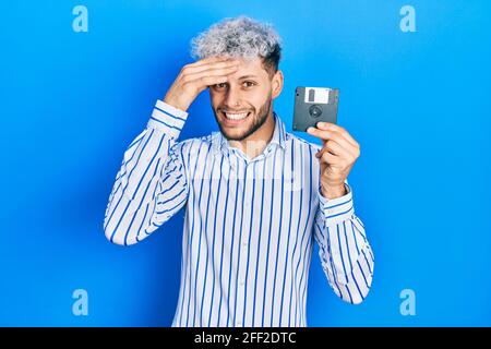 Giovane uomo ispanico con moderni capelli tinti tenendo floppy disk stressato e frustrato con la mano sulla testa, sorpreso e arrabbiato viso Foto Stock