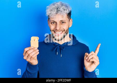 Giovane uomo ispanico con moderni capelli tinti che tengono gli auricolari wireless sorridendo felice puntando con mano e dito a lato Foto Stock