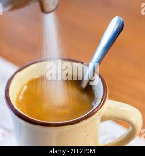 Lo zucchero viene versato da un distributore di zucchero in una tazza di caffè calda al ristorante al mattino. L'impugnatura del cucchiaio fuoriesce dalla tazza. Foto Stock