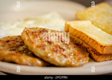 Un piatto in un ristorante per la colazione che include 2 polpettine di salsiccia di maiale, due fette di pane tostato bianco e 2 sopra uova facili sfocate sullo sfondo. Piastra bianca. Foto Stock