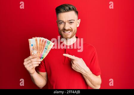 Giovane uomo rosso che tiene le banconote del peso filippino sorridente che indica felice con mano e dito Foto Stock