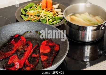 Padella con pepe rosso arrosto, pentola con asparagi bollenti e piatto con verdure tagliate su stufa in ceramica calda. Preparazione di un pasto vegetariano. Foto Stock