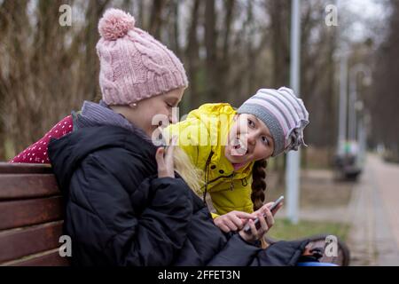 due ragazze sono sedute in un parco su una panchina con i telefoni nelle loro mani, gli scooter sono in piedi nelle vicinanze. concetto di dipendenza dei bambini dallo smartphon Foto Stock