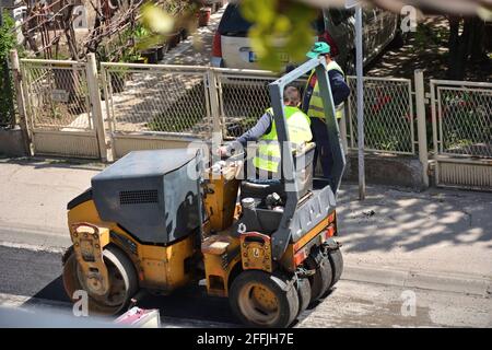 NIS, Serbia - 24 aprile 2021: Compattatore a rulli gommato piccolo su asfalto lavori di riparazione stradale con lavoratori in uniforme verde Foto Stock