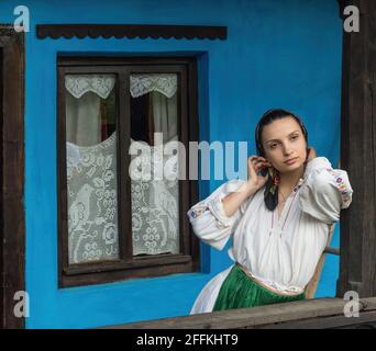 Salaj, Transilvania, Romania-14 maggio 2018: Bella giovane ragazza in costume tradizionale rumeno folk seduta sulla terrazza di una piccola casa di charme Foto Stock