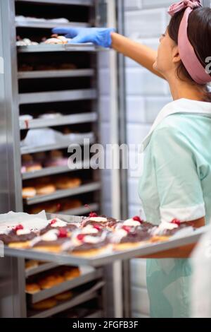 Una giovane piccola impresa donna proprietario sta prendendo le ciambelle deliziose fatte a mano fuori da un owen in un'atmosfera di lavoro in un laboratorio caramelle. Pasticceria, dessert, Foto Stock