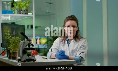 POV della donna botanista in cappotto bianco che ascolta il team chimico durante la riunione on-line di video mentre si siede in laboratorio farmaceutico. Team di specialisti che ricercano la mutazione genetica che sviluppa il test di biologia degli OGM del dna Foto Stock