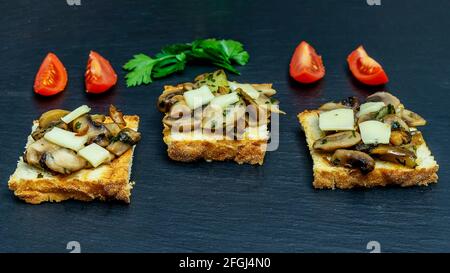 Tre fette di pane tostato con funghi champignon, prezzemolo, formaggio emmental e pomodori ciliegia rossi a fette, su piastra di ardesia Foto Stock